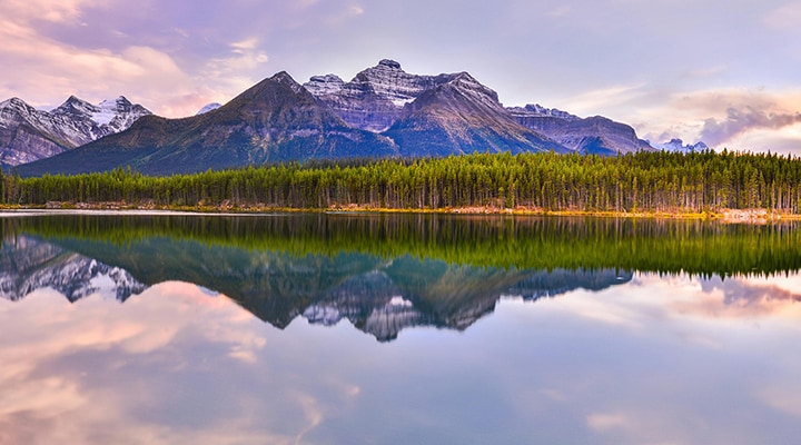 Mountain range reflected in a calm lake at sunrise, with evergreen forest along the shoreline and soft pastel sky.